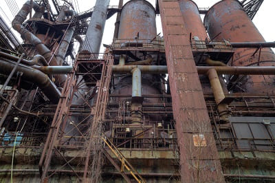 Abandoned blast furnace with vegetation growing through rusted steelworks structure — derelict zombie steel mill kept alive by state subsidy